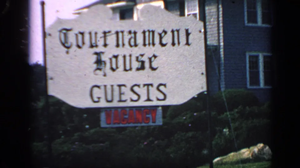 Close view of the Tournament House guest sign and red vacancy notice, with bushes and a New England-style house in the background.