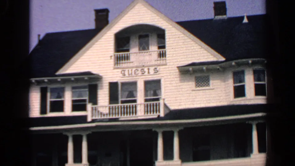 Massachusetts guesthouse exterior in 1968 footage, showing a white clapboard facade, porch columns, balcony, windows, and guests lettering.