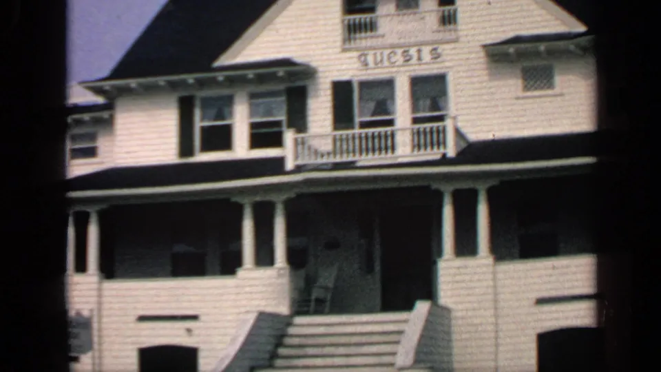 1968 Massachusetts home-movie view of a white house exterior with central steps, porch columns, balcony railing, and GUESTS sign.