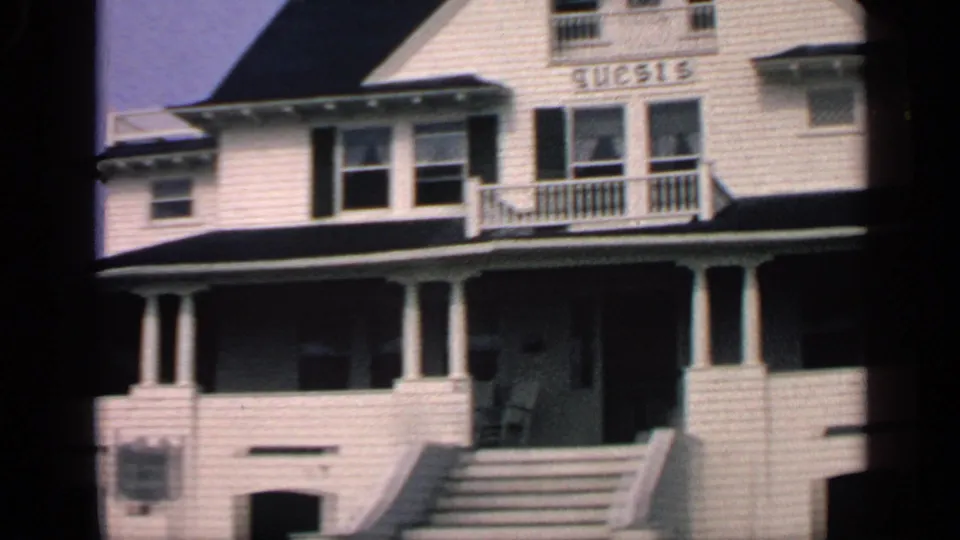 1968 Massachusetts 8mm frame of a white shingled guest-house exterior with front stairs, shaded porch, balcony doors, and sign lettering.