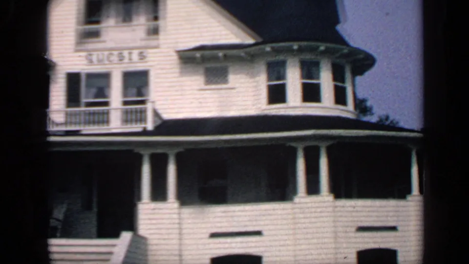 1968 Massachusetts home-movie frame showing the right side of a white shingled house with bay windows, porch columns, balcony, and GUESTS sign.
