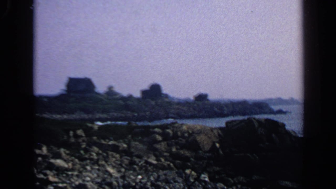1968 Massachusetts coastal view with dark rocks, ocean water, distant houses, and soft 8mm framing.