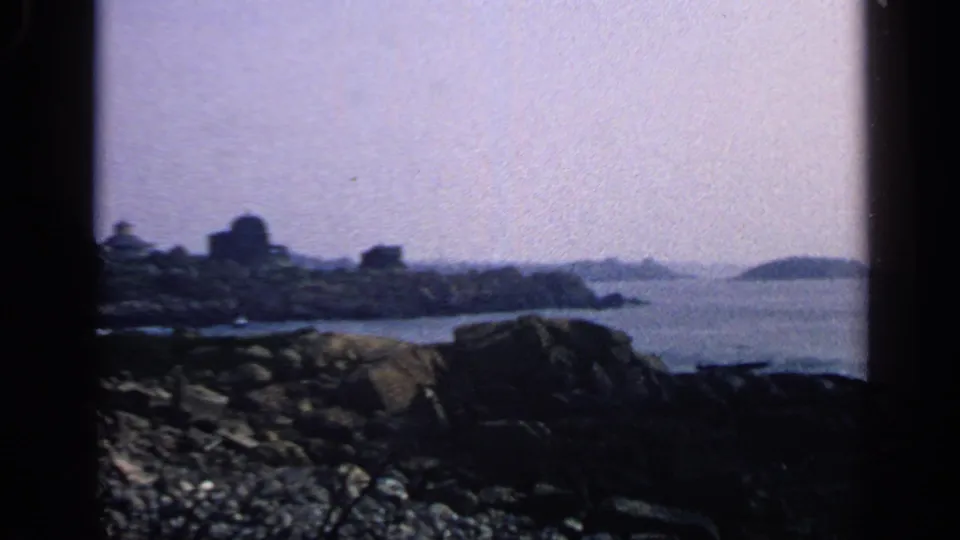 Home-movie view of a Massachusetts rocky shoreline with water, distant coastal buildings, and a low island beyond.