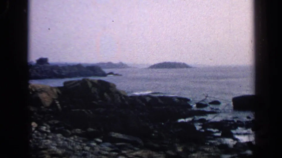 1968 Massachusetts shoreline frame with dark coastal rocks, open water, distant land, and a small offshore island.