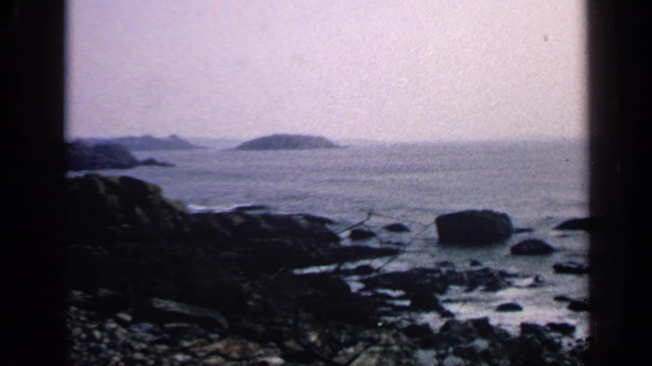 Rocky shoreline with dark boulders, calm sea, hazy sky, and distant low island forms.