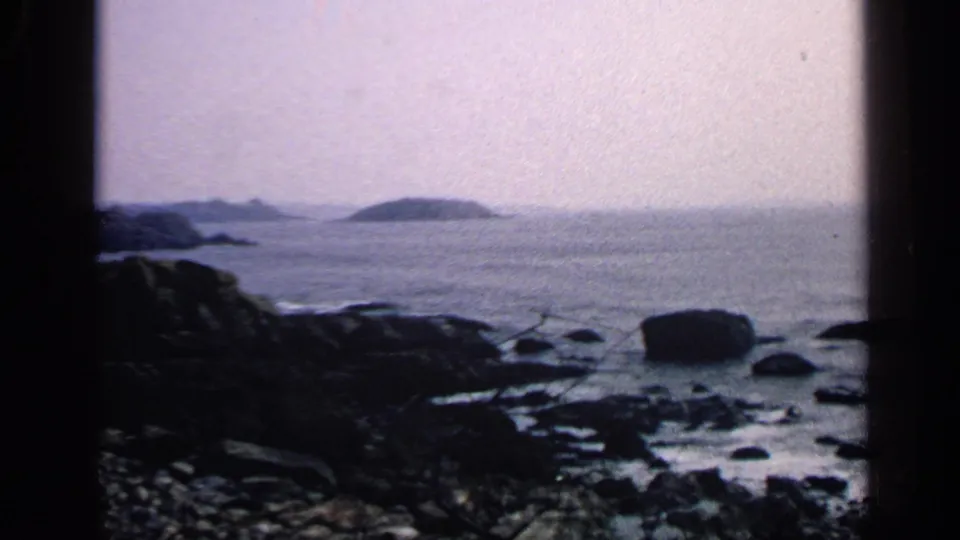 1968 Massachusetts 8mm frame showing a rocky coast, dark foreground rocks, calm water, hazy horizon, and distant island shapes.