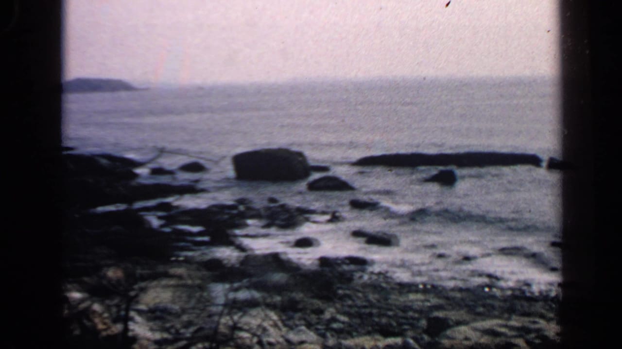 1968 Massachusetts coastal home-movie frame with calm water, scattered rocks, a large boulder, distant land, and dark window-like framing.