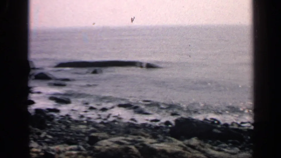 1968 Massachusetts 8mm view of a pebbled rocky shore, small waves, calm sea, and low offshore rock shelf under a pale sky.