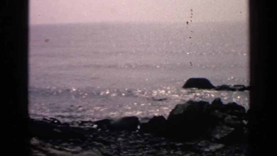 1968 Massachusetts coastal frame with shimmering water, dark rocks in the foreground, a boulder silhouette, hazy horizon, and visible film specks.