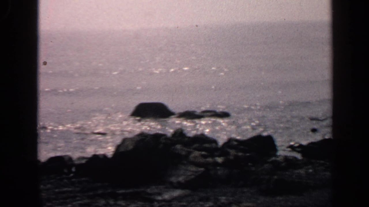 1968 Massachusetts home-movie frame of a rocky shore and shimmering water viewed through dark foreground framing.