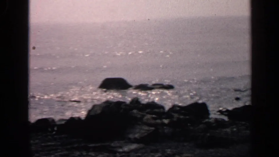 1968 Massachusetts home-movie frame of a rocky shore and shimmering water viewed through dark foreground framing.