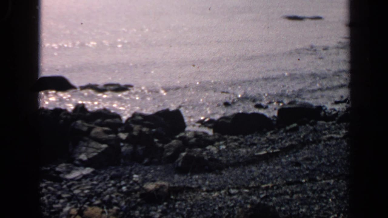 Massachusetts coastal view with rocks, pebbles, and reflective water, partially blocked by dark foreground edges.
