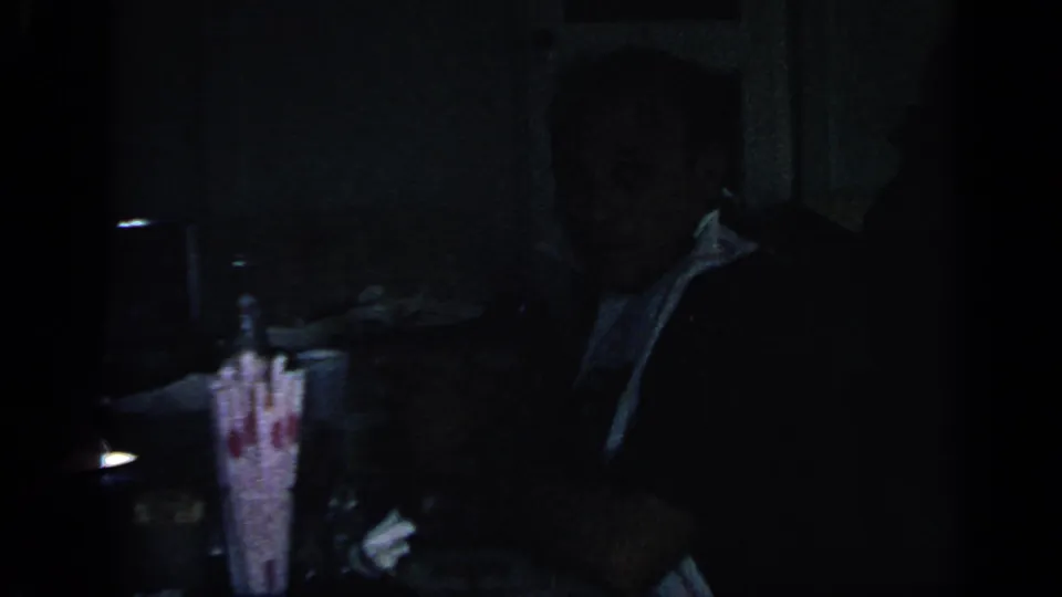 Dim 1968 home-movie interior showing an adult seated near a table, with details partly lost in shadow.
