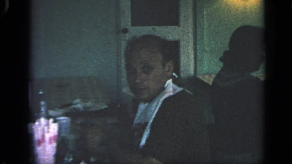 Dim Massachusetts home-movie interior showing a suited man at a table and another adult in side silhouette.