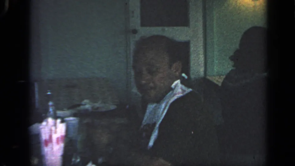 1968 Massachusetts home-movie frame of an older man seated in a dim restaurant with a white bib, with tableware nearby.