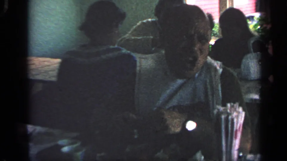 1968 Massachusetts home-movie frame showing seated diners in a low-lit restaurant, with table items and a window area visible.