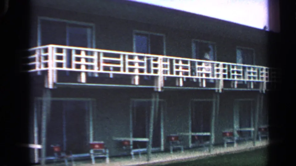 1968 Massachusetts frame of a two-story motel exterior with long white balcony railings, room doors, lawn, and chairs outside rooms.