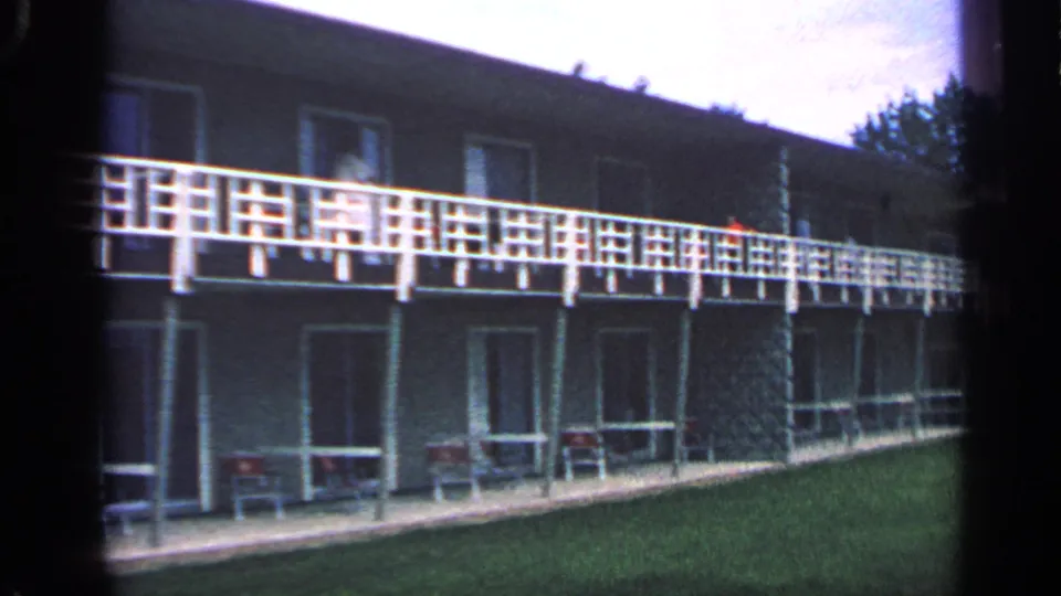 1968 Massachusetts home-movie frame showing a longer view of the motel facade, balcony railings, room fronts, lawn, and trees beyond.