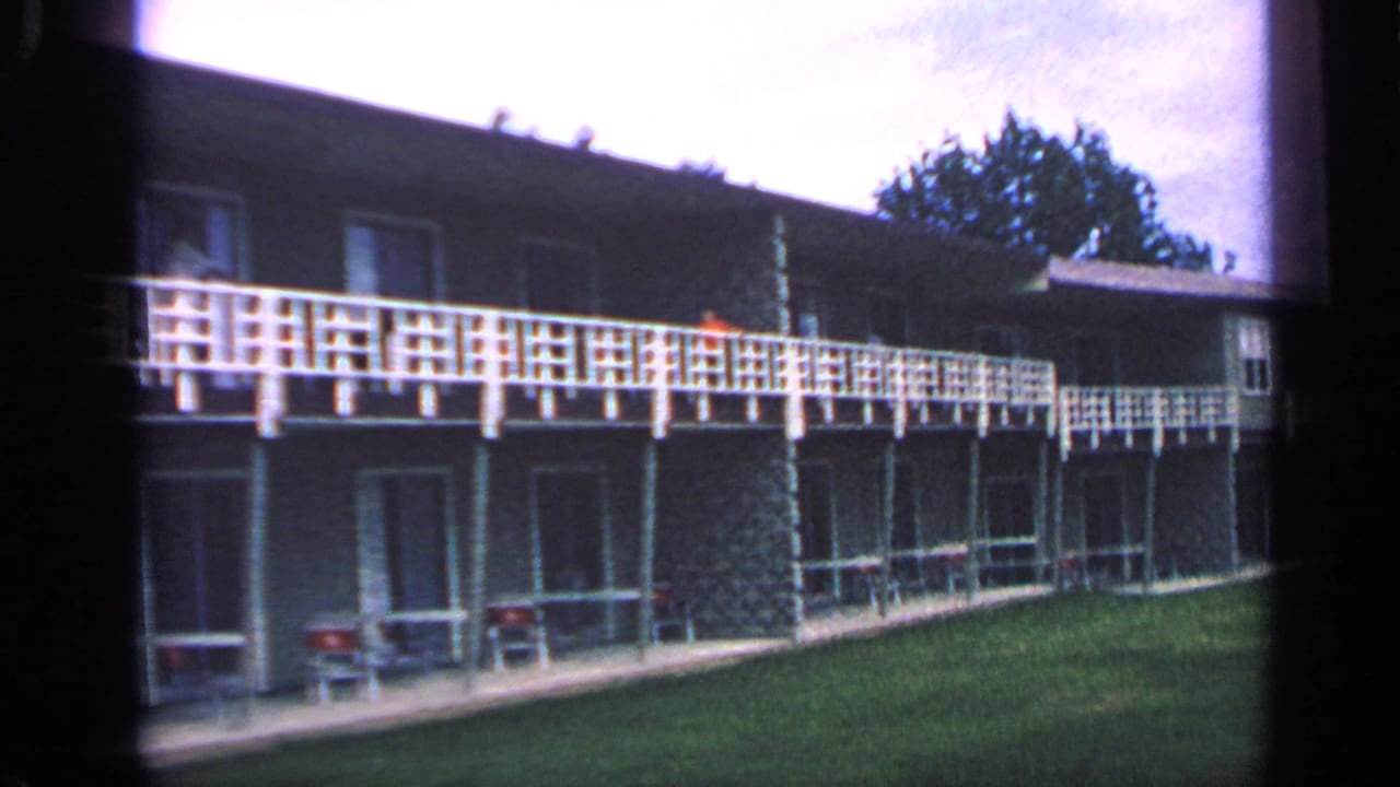 Two-story apartment building with white balcony railing, lawn, trees, and overcast sky.