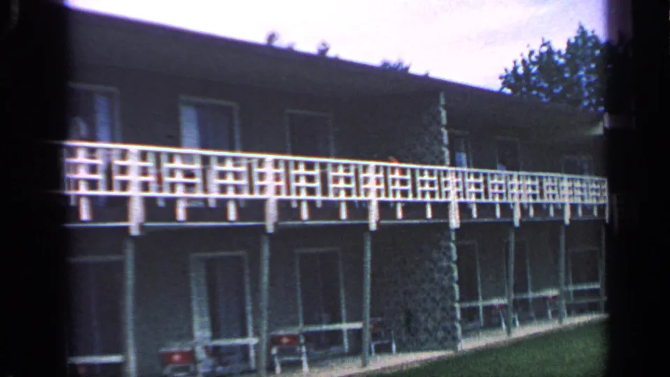 1968 Massachusetts apartment exterior with white second-floor balcony rail, shaded lower walkway, chairs, lawn, and pale sky.