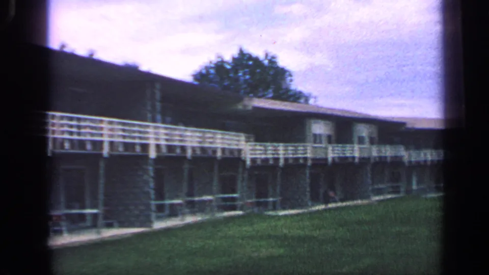 Wide 1968 Massachusetts 8mm view looking along the apartment row, with white balcony rail, lawn foreground, trees, and cloudy sky.