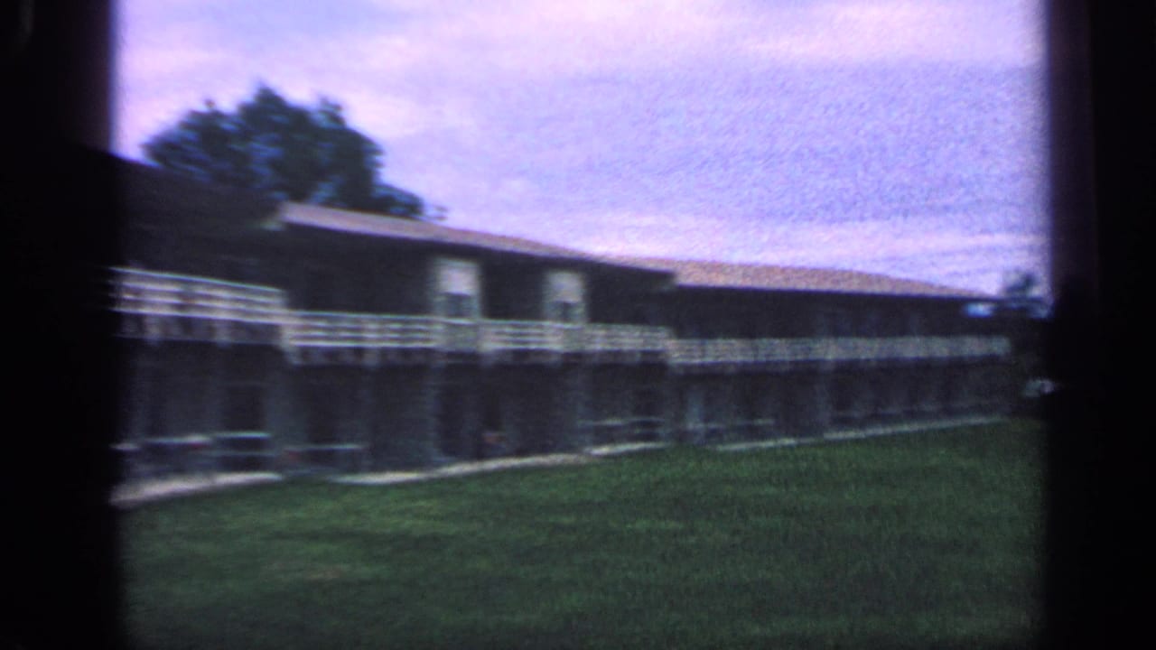 Low apartment building with white railings beside a grassy lawn under a cloudy sky.