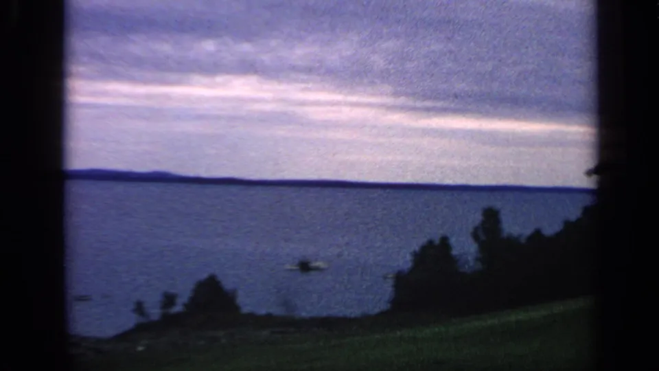 1968 Massachusetts lake view with a low horizon, cloudy sky, dark shoreline trees, grassy foreground, and a small boat on the water.