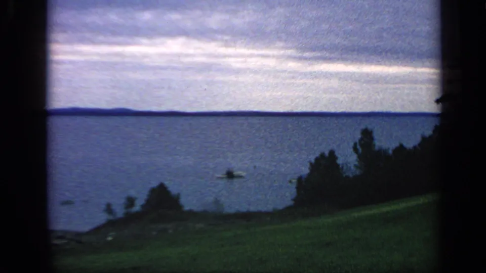 1968 Massachusetts 8mm frame showing a calm lake beyond a grassy slope, with trees, small boats, distant shoreline, and heavy cloud cover.
