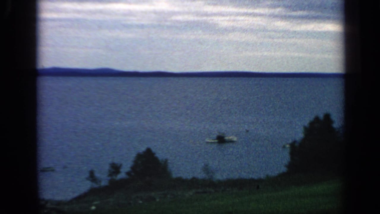 Lake view with distant hills, gray sky, shoreline trees, grass, and a small boat on the water.