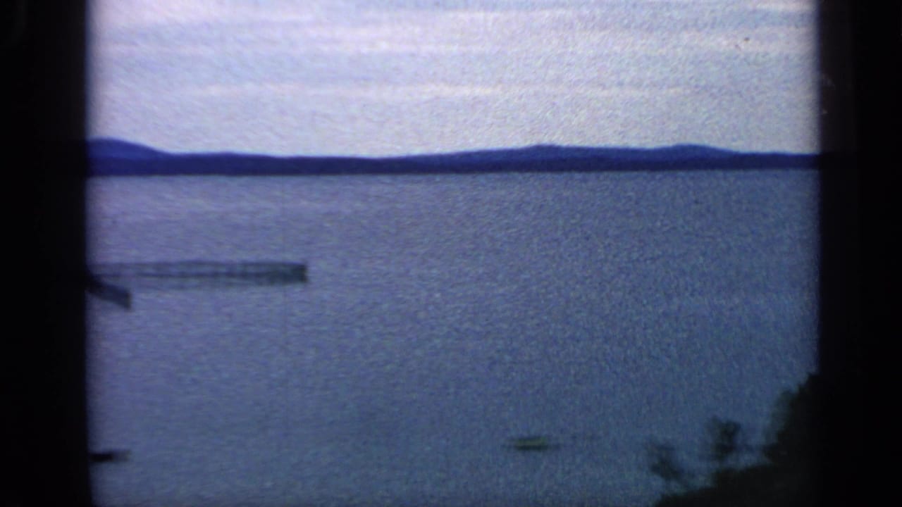 Open lake with distant hills, gray sky, a small boat, and a narrow dock-like shape on the water.