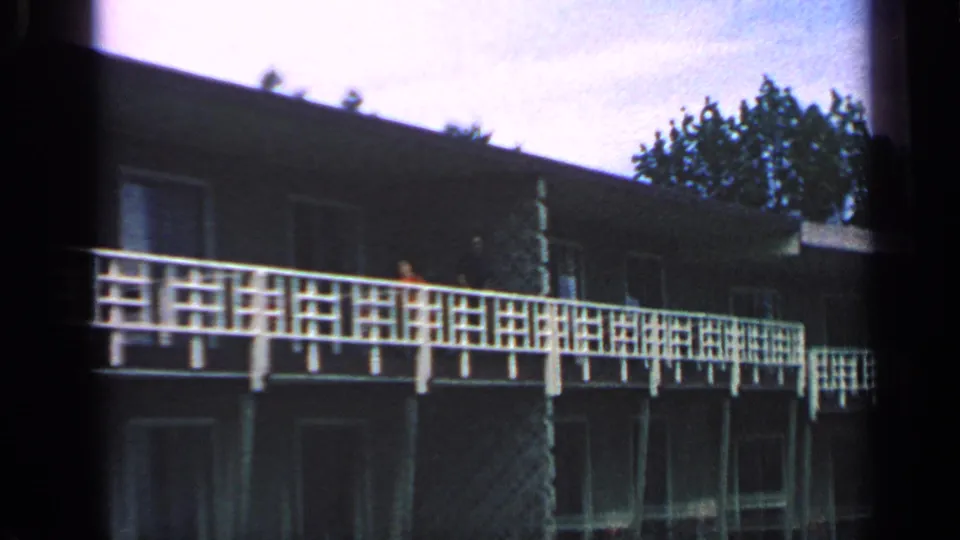 1968 Massachusetts 8mm frame of a waterfront building with a long white balcony rail, dark exterior, windows, roofline, and nearby trees.