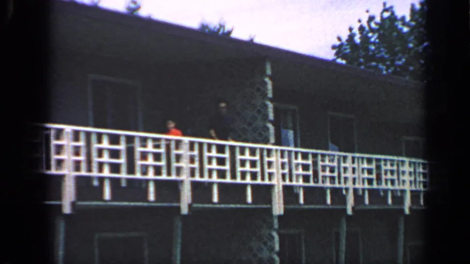 1968 Massachusetts home-movie frame showing a dark building facade, white balcony railing, and tiny figures on an elevated walkway.