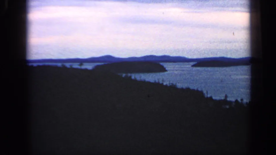 1968 Massachusetts 8mm frame of a wide water landscape with silhouetted woods, small islands, and hazy hills in the distance.