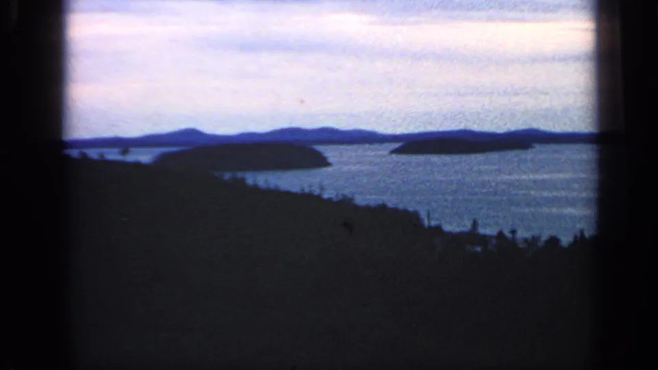 1968 8mm Massachusetts coastal view showing calm water, low islands, distant hills, and silhouetted trees in the foreground.