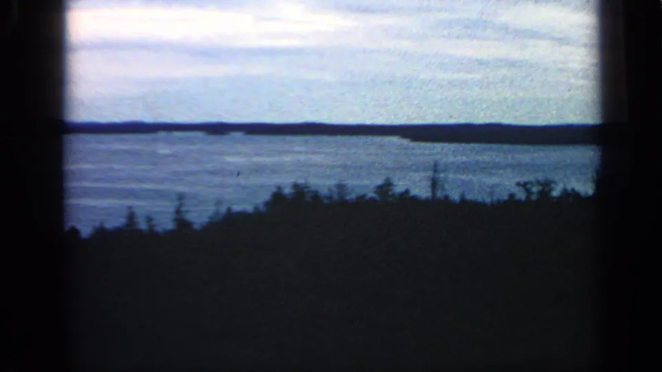 1968 coastal home-movie view in Massachusetts showing open water, a low island, distant shore, and shadowed foreground vegetation.