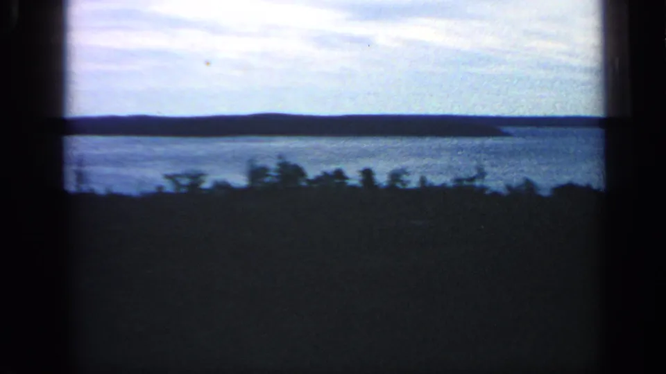 Blue-toned 1968 8mm frame of a Massachusetts coastal panorama with calm water, long low landform, and dark foreground trees.