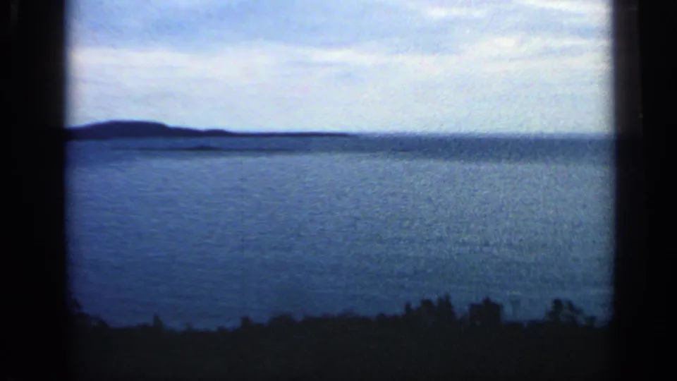 1968 Massachusetts 8mm still of open water, faint horizon landforms, pale sky, and a dark wooded foreground edge.