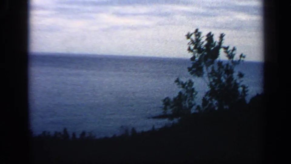 1968 Massachusetts 8mm frame showing blue water, pale sky, dark shoreline, and a tree silhouette at right.