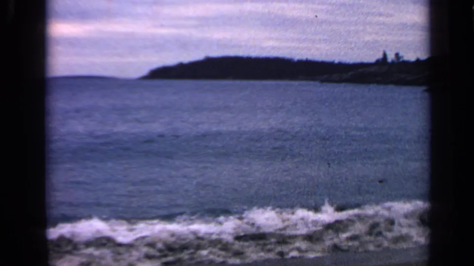 1968 Massachusetts coastal home-movie frame with white surf in the foreground and a low wooded shoreline under a pale sky.