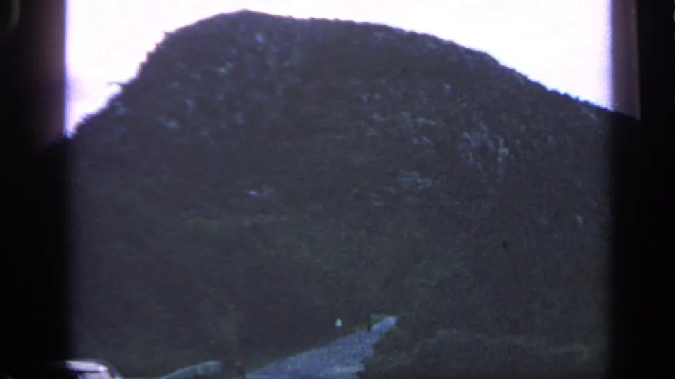 1968 Massachusetts 8mm frame showing a dark tree-covered coastal hill, low vegetation, and a narrow path or rocky strip below.