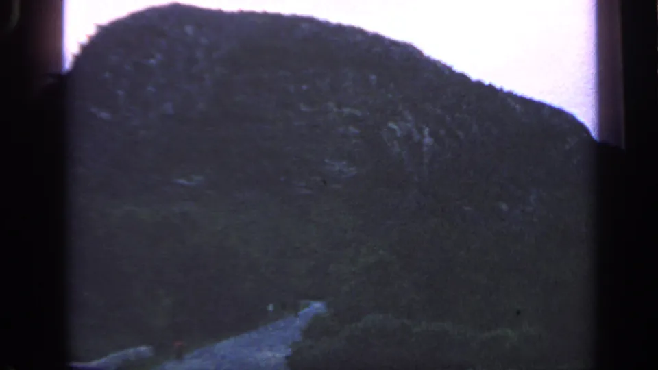 1968 Massachusetts coastal frame with a shadowed wooded hill, faint rocky texture, low vegetation, and soft overcast light.