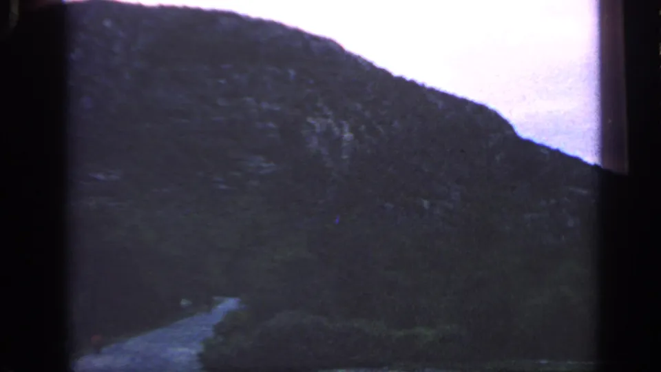 1968 Massachusetts 8mm frame showing a dark coastal hillside, a faint winding path, and water beneath a soft evening sky.