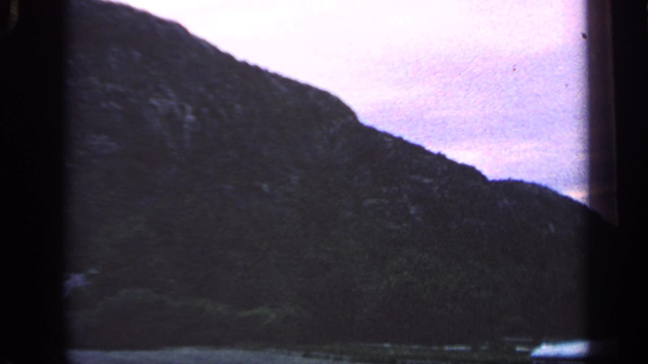 1968 Massachusetts coastal view with a shadowed hill, narrow waterline, and soft purple sky in an 8mm home-movie frame.