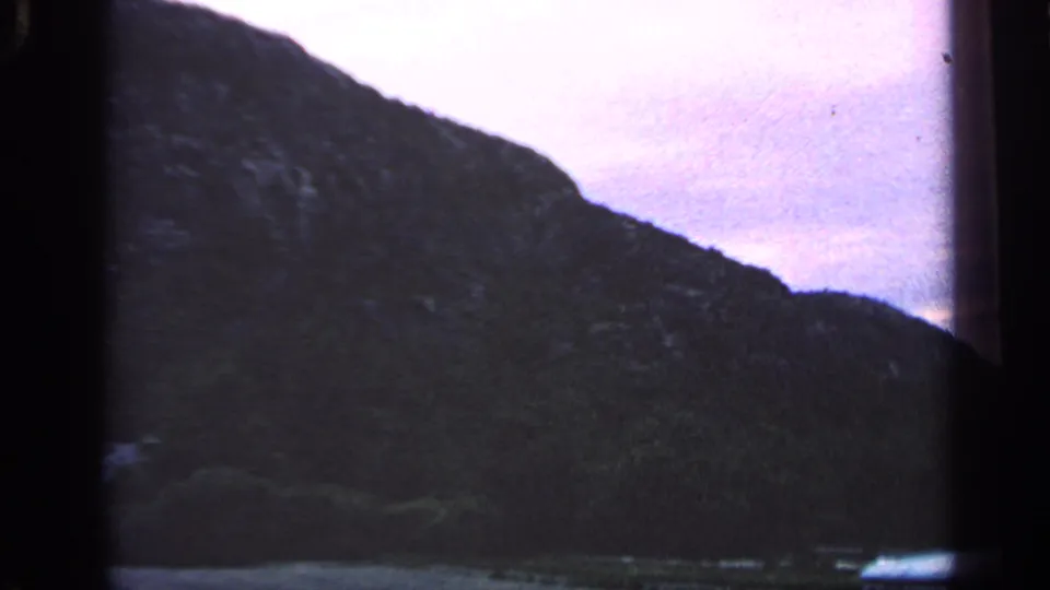 1968 Massachusetts coastal view with a shadowed hill, narrow waterline, and soft purple sky in an 8mm home-movie frame.
