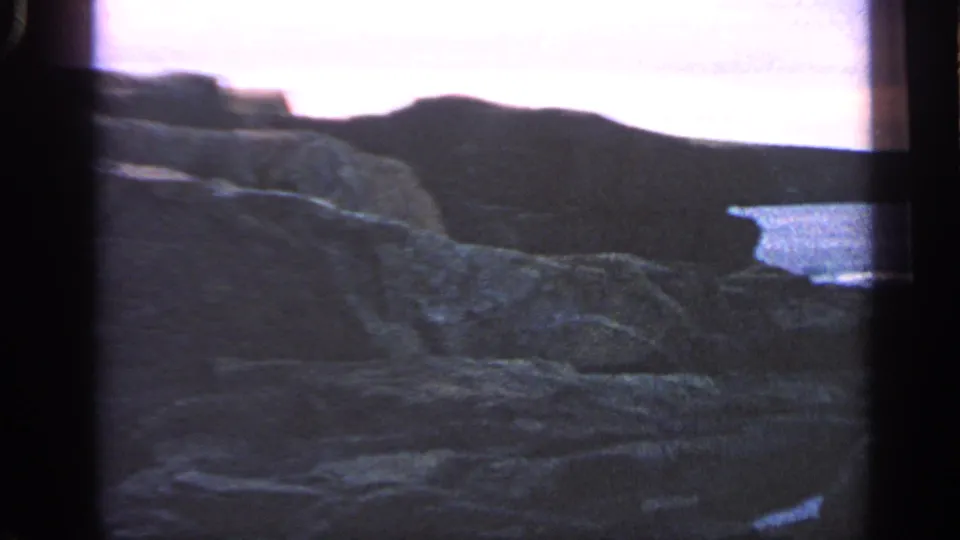 1968 Massachusetts 8mm view across uneven coastal rocks toward low cliffs, calm water, and a bright dusk sky.