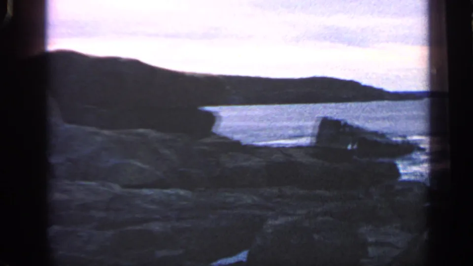 1968 Massachusetts coastal frame with jagged rocks in front, calm sea beyond, and a dark headland under a pale evening sky.
