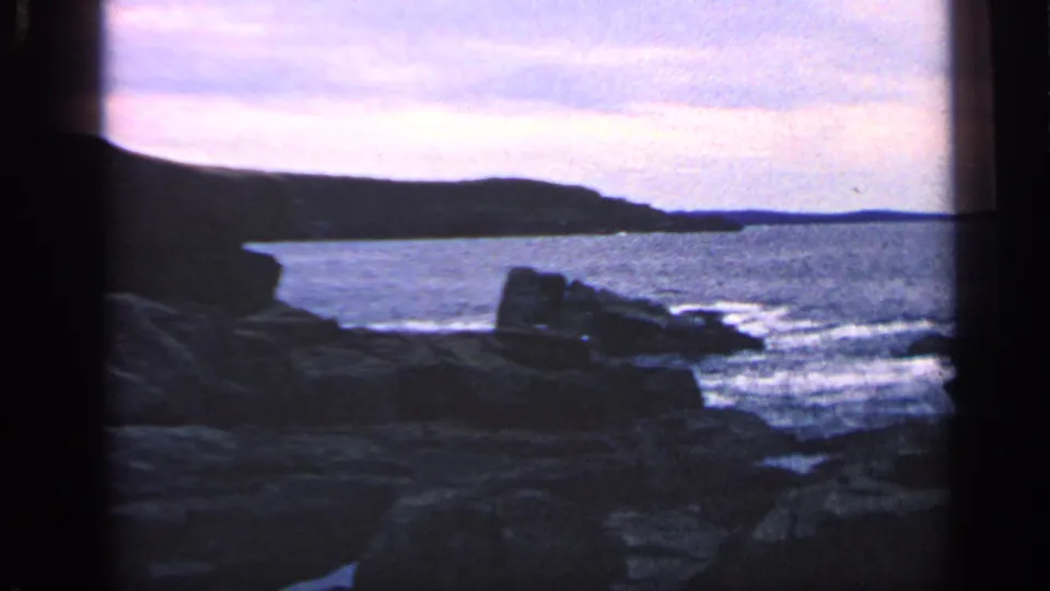 1968 Massachusetts 8mm frame of a rocky coastal cove with surf, distant headland, and pink-purple twilight sky.