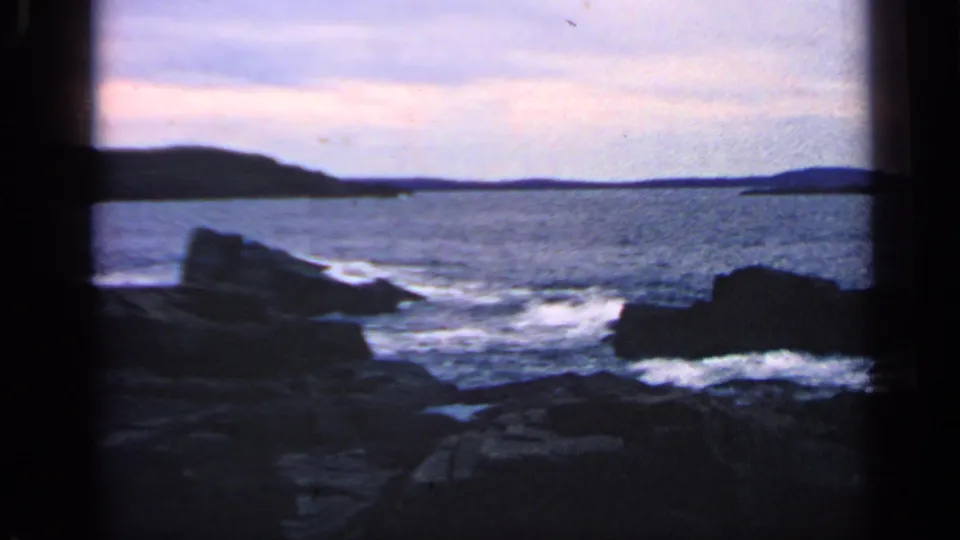 1968 Massachusetts coastal home-movie frame showing waves around jagged rocks beneath a dim twilight sky.