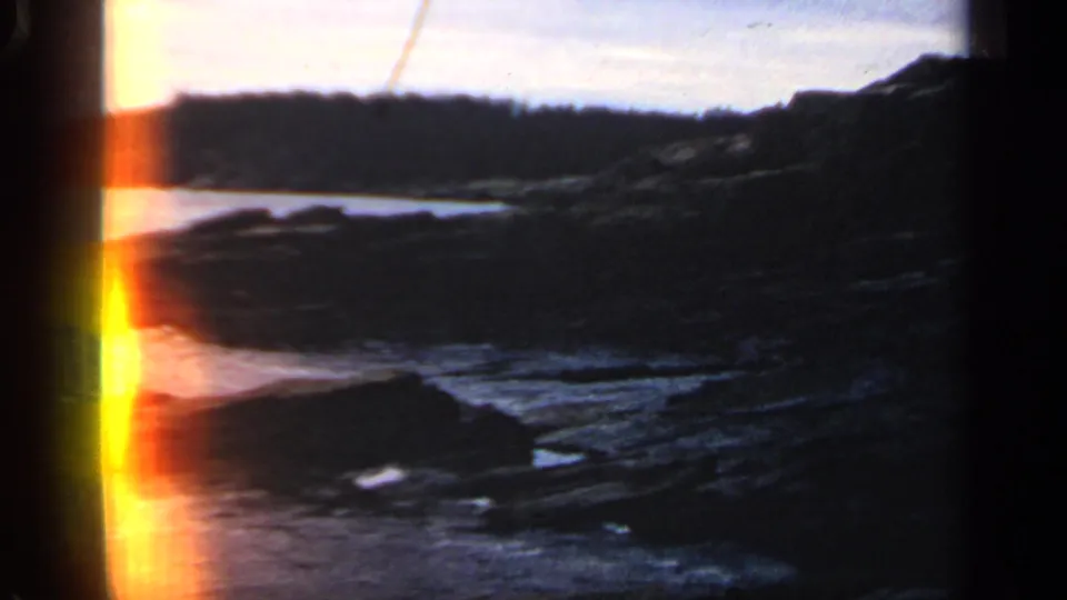 1968 Massachusetts 8mm frame of a shadowed rocky cove, low surf, wooded shoreline, and visible film-edge light leak.