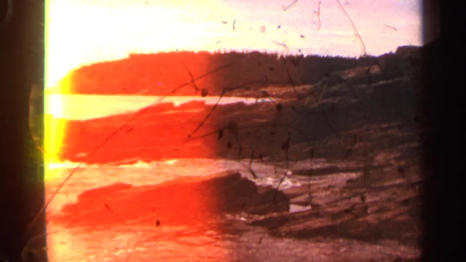 1968 Massachusetts 8mm frame with strong red-orange exposure flare over surf, rocky shore, distant trees, and film scratches.
